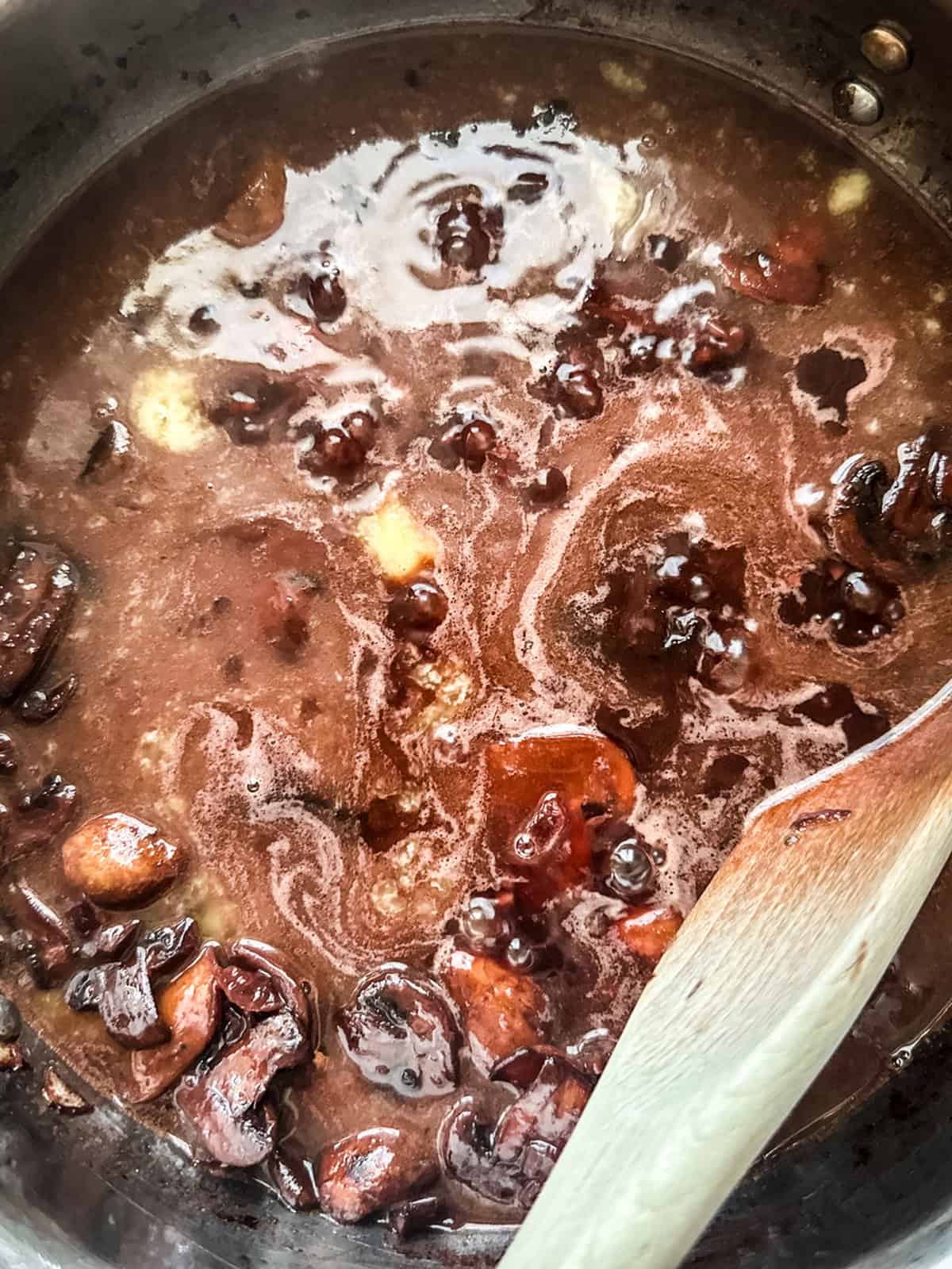 Mushrooms in red wine sauce with butter and a wooden spoon in a skillet.