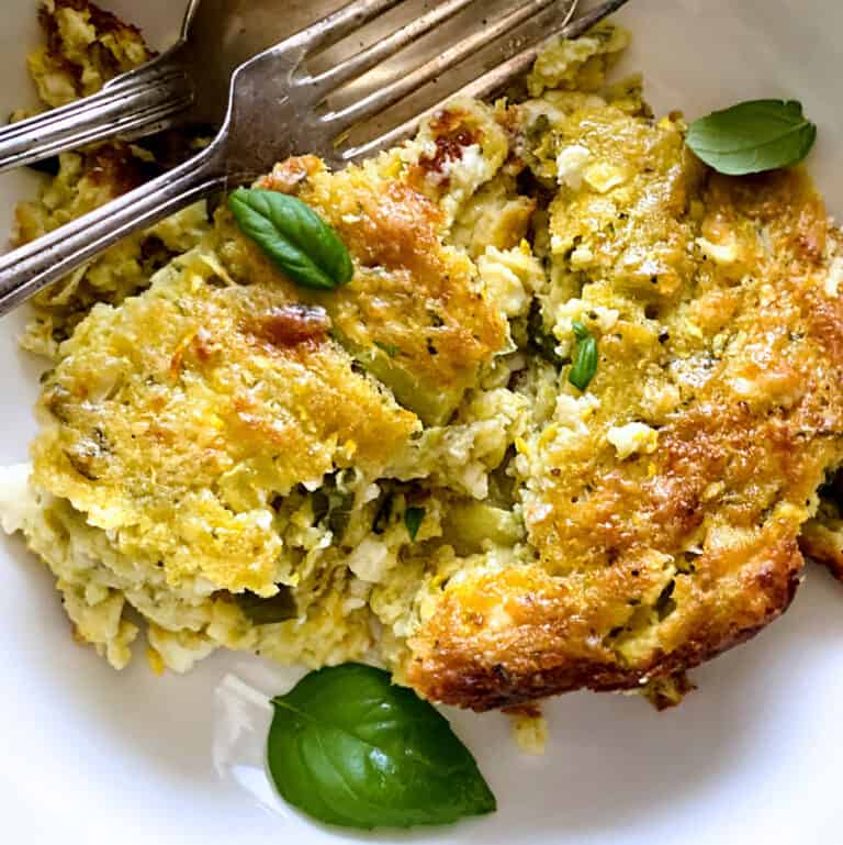 A piece of crusted zucchini pie on a plate with a fork and spoon. Some basil leaves on the pie.