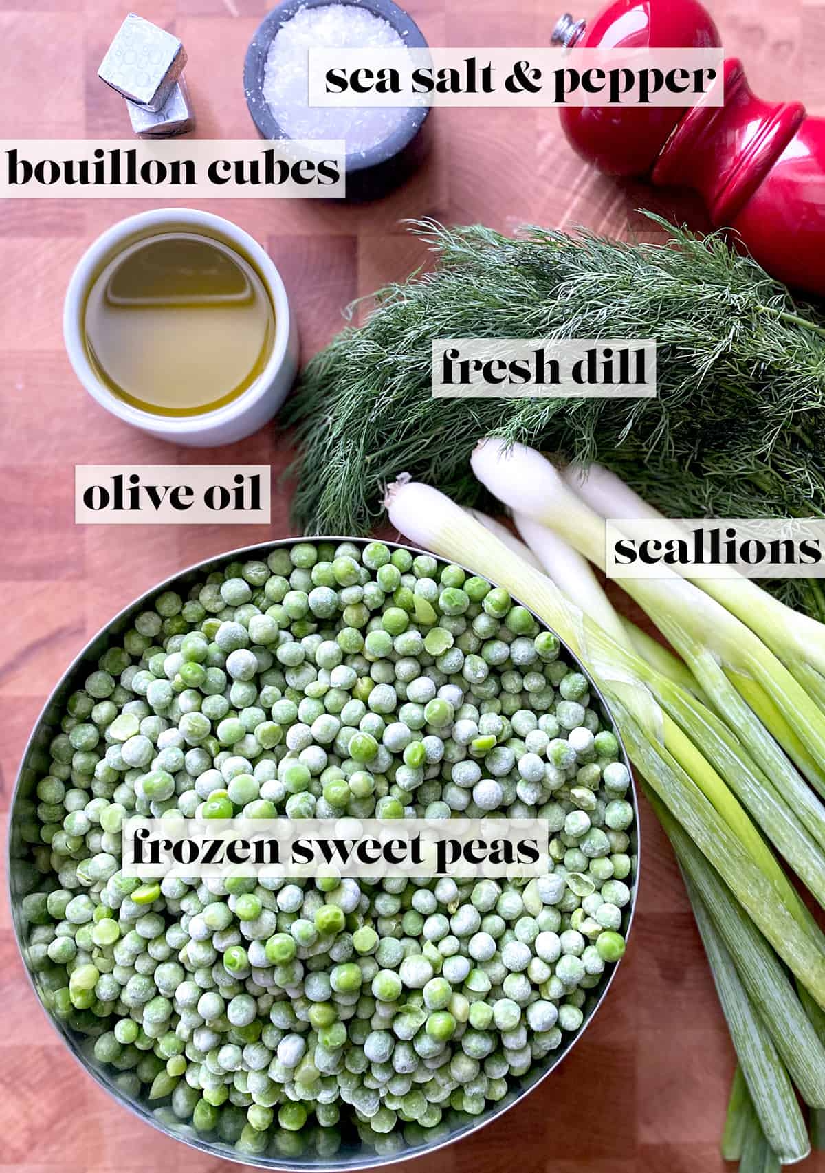 On aa butcher block, two bouillon cubes, a small bowl with sea salt, a red pepper mill, a small container with olive oil, a bunch of fresh dill, five fresh scallions and a large bowl with frozen peas.