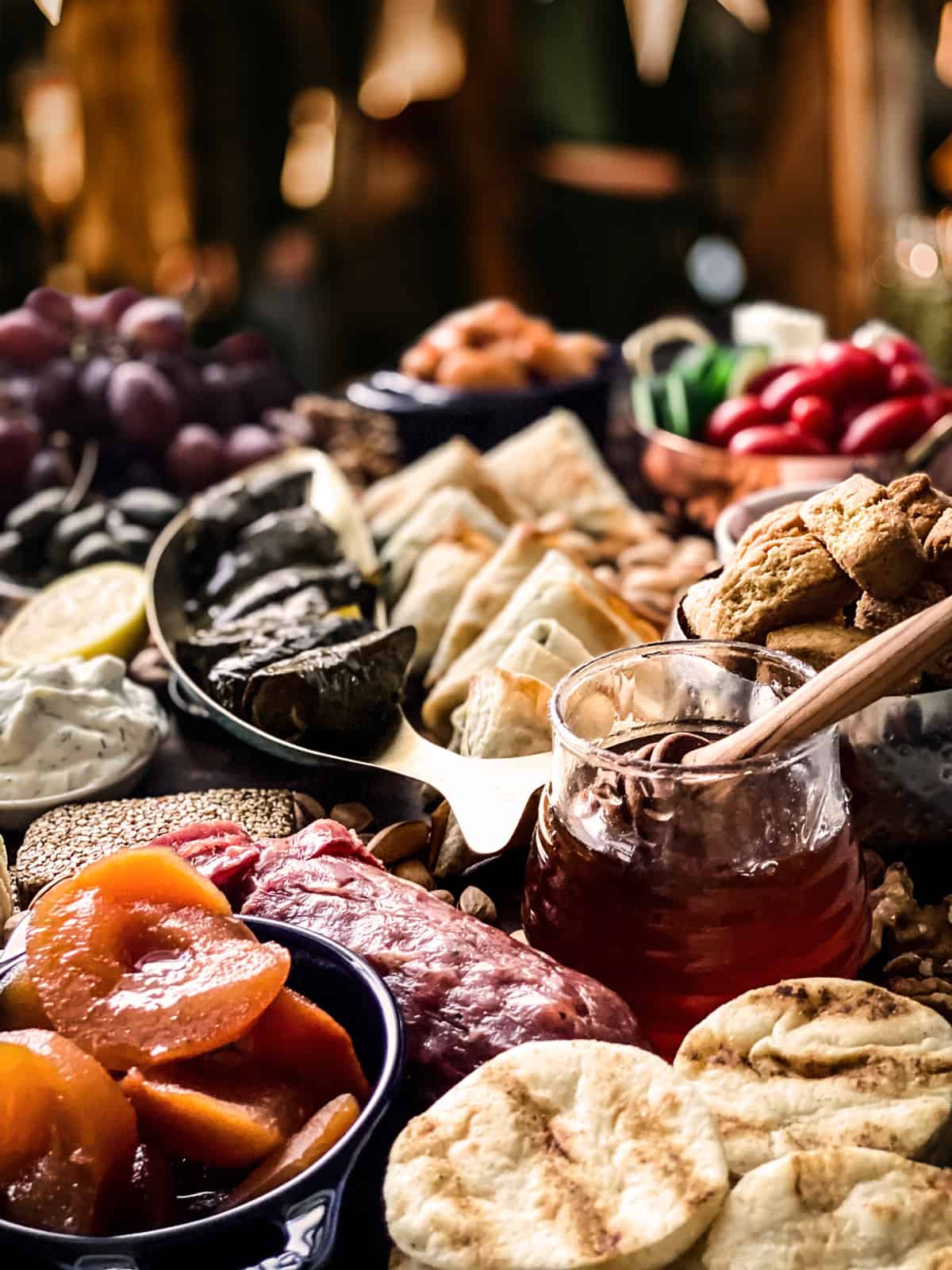 Grapes, walnuts, a small bowl with beans, a fish shaped bowl with dolmades, half a lemon, sliced salami, a pasteli bar, a pot with preserved apricots, a pot of honey, a small pot with fig preserves, some pistachios, round wafers, sliced cheese, and tzatziki dip.