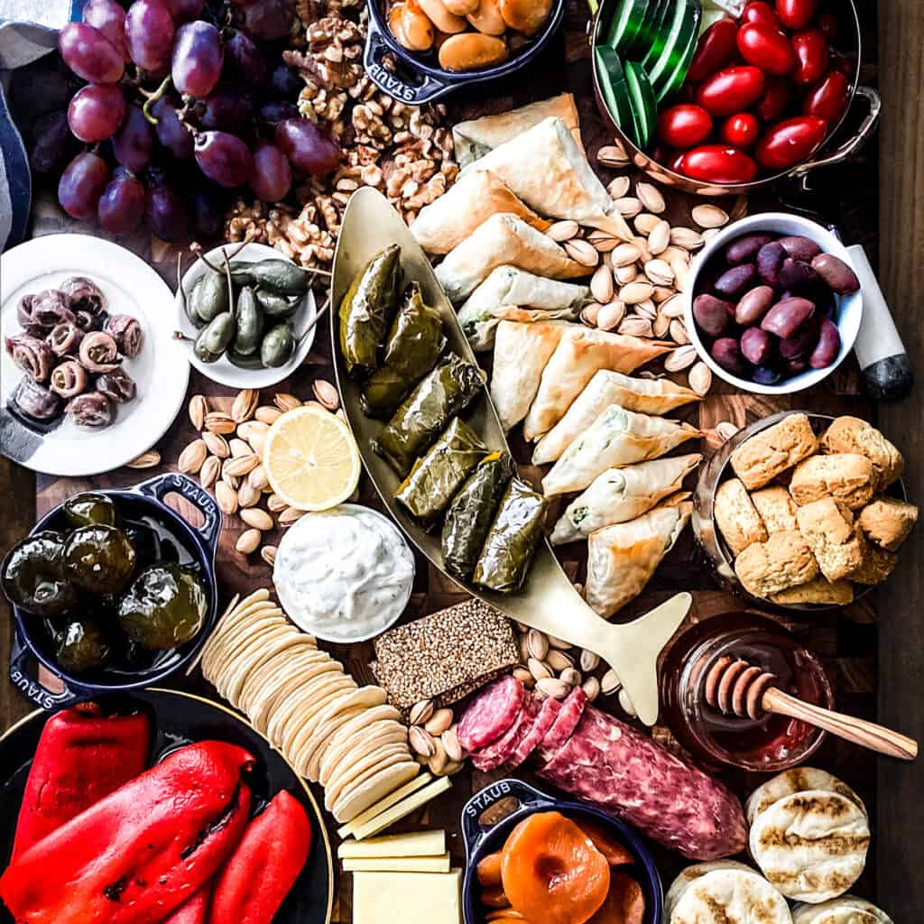 A small pot with fig preserves, some pistachios, round wafers, a side view of roasted peppers on a plate and side view of tzatziki dip and anchovies.