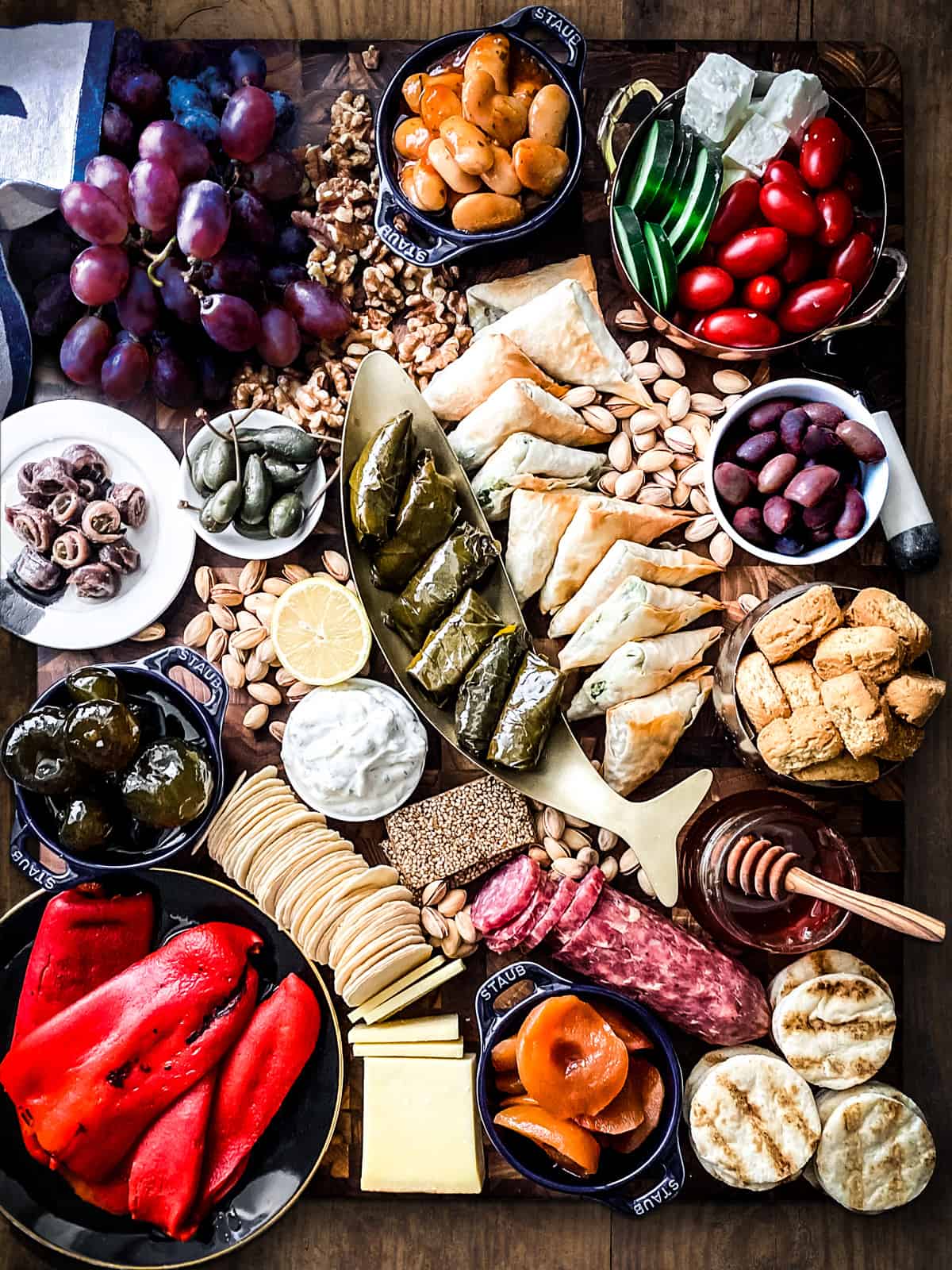 Grapes, walnuts, a small bowl with beans, a fish shaped bowl with dolmades, a salami, some pastel, a pot with preserved apricots, a pot of honey, a small pot with fig preserves, some pistachios, round wafers, a side view of roasted peppers on a plate and side view of tzatziki dip and anchovies.