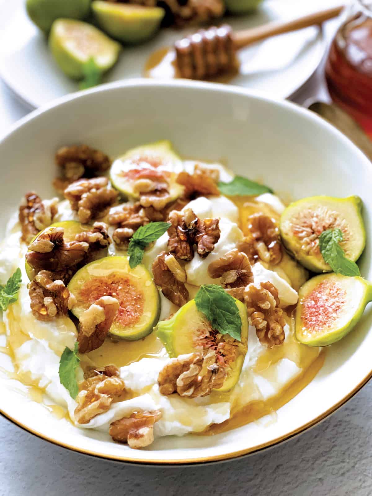 A plate with yogurt, sliced figs, walnuts and honey and a spoon. At the back a plate with figs and nuts, a honey dripper and a pot of honey.