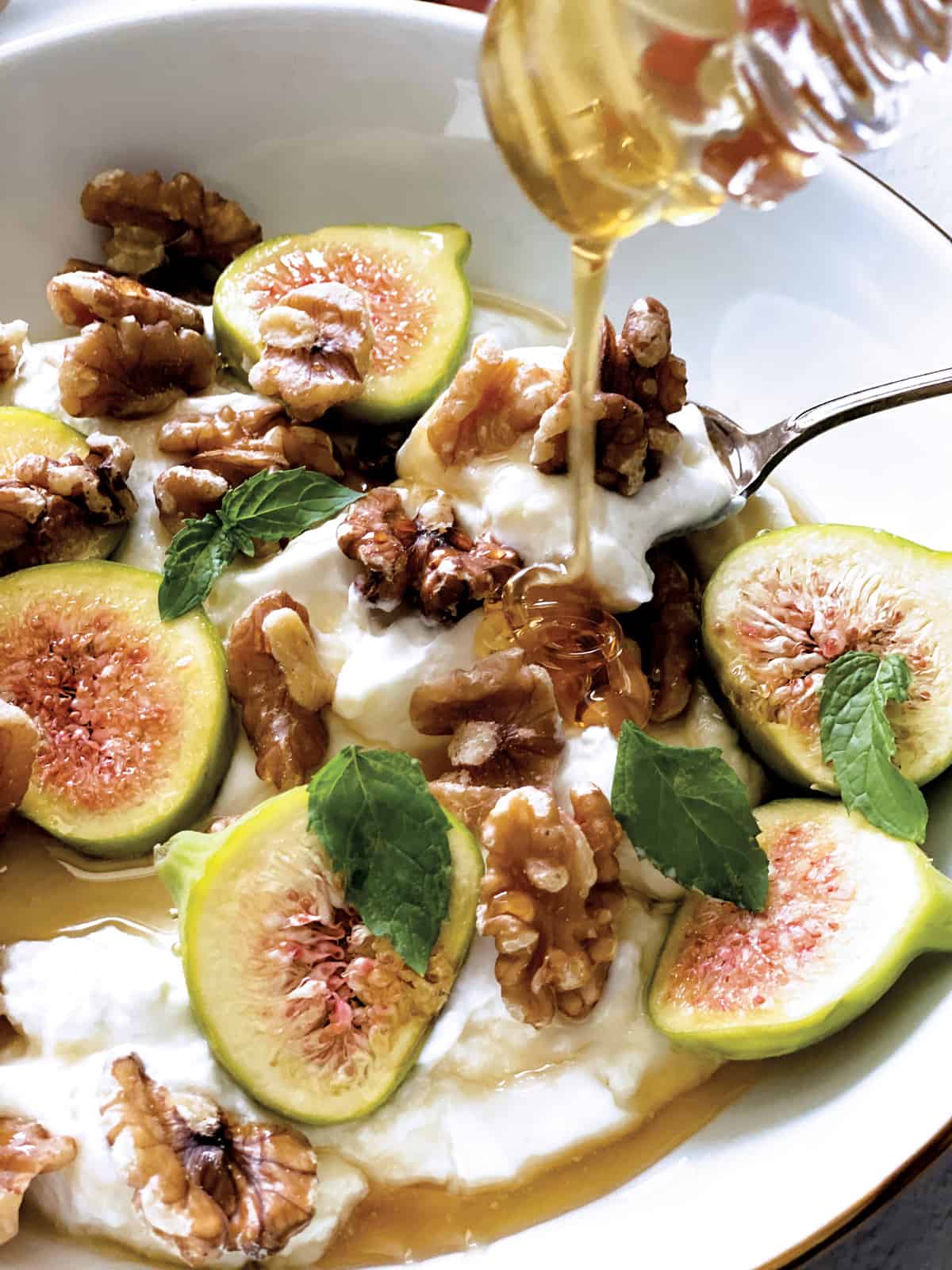 Close up of yogurt, figs and walnuts and a honey dipper drizzling honey.