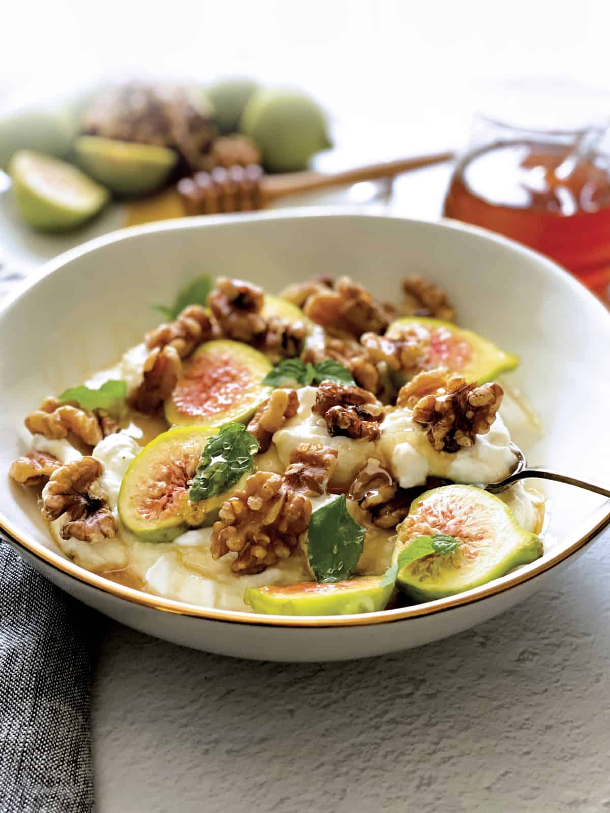 A plate with yogurt, sliced figs, walnuts and honey and a spoon. At the back a plate with figs and nuts, a honey dripper and a pot of honey.