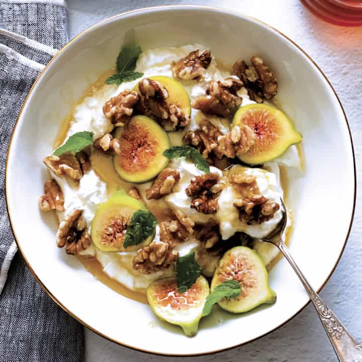 A plate with yogurt, sliced figs, walnuts and honey and a spoon. At the back a plate with figs and nuts.