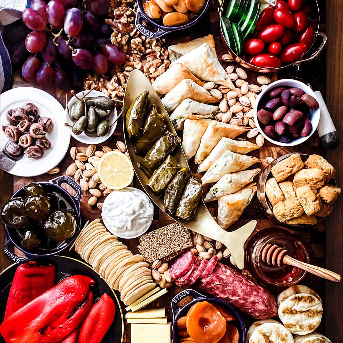 Overhead Greek grazing board with dolmadakia in a fish-shaped dish, spanakopitakia, tzatziki, feta, olives, grapes, salami, and grilled pita