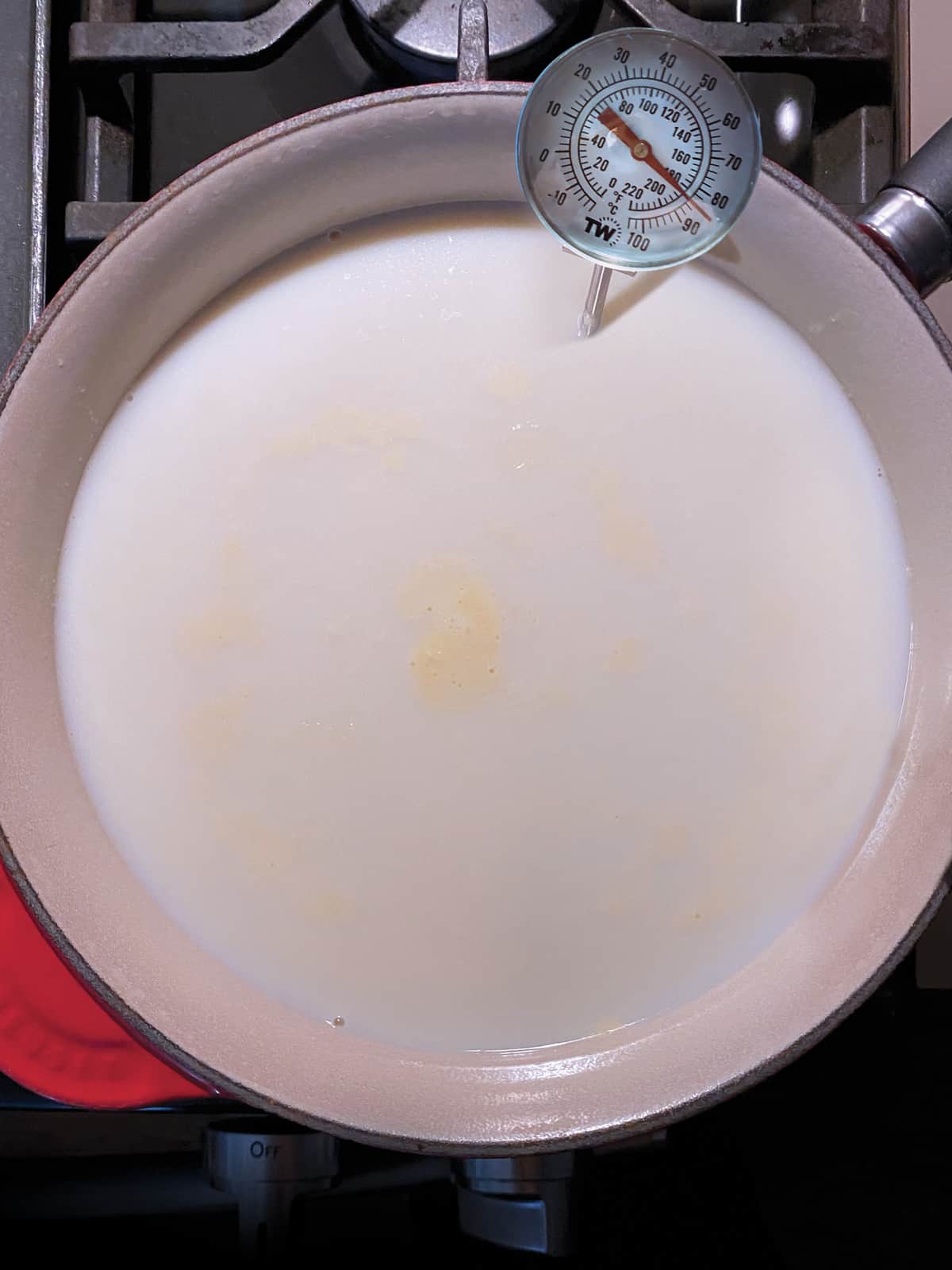 A sauce pan on the stove full with milk and a milk thermometer.
