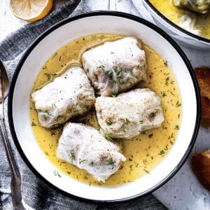 A plate with stuffed cabbage rolls, lahanodolmades, a piece of bead at the top and right, part of the pot is showing and part of utensils with a cloth napkin, all on a white table.