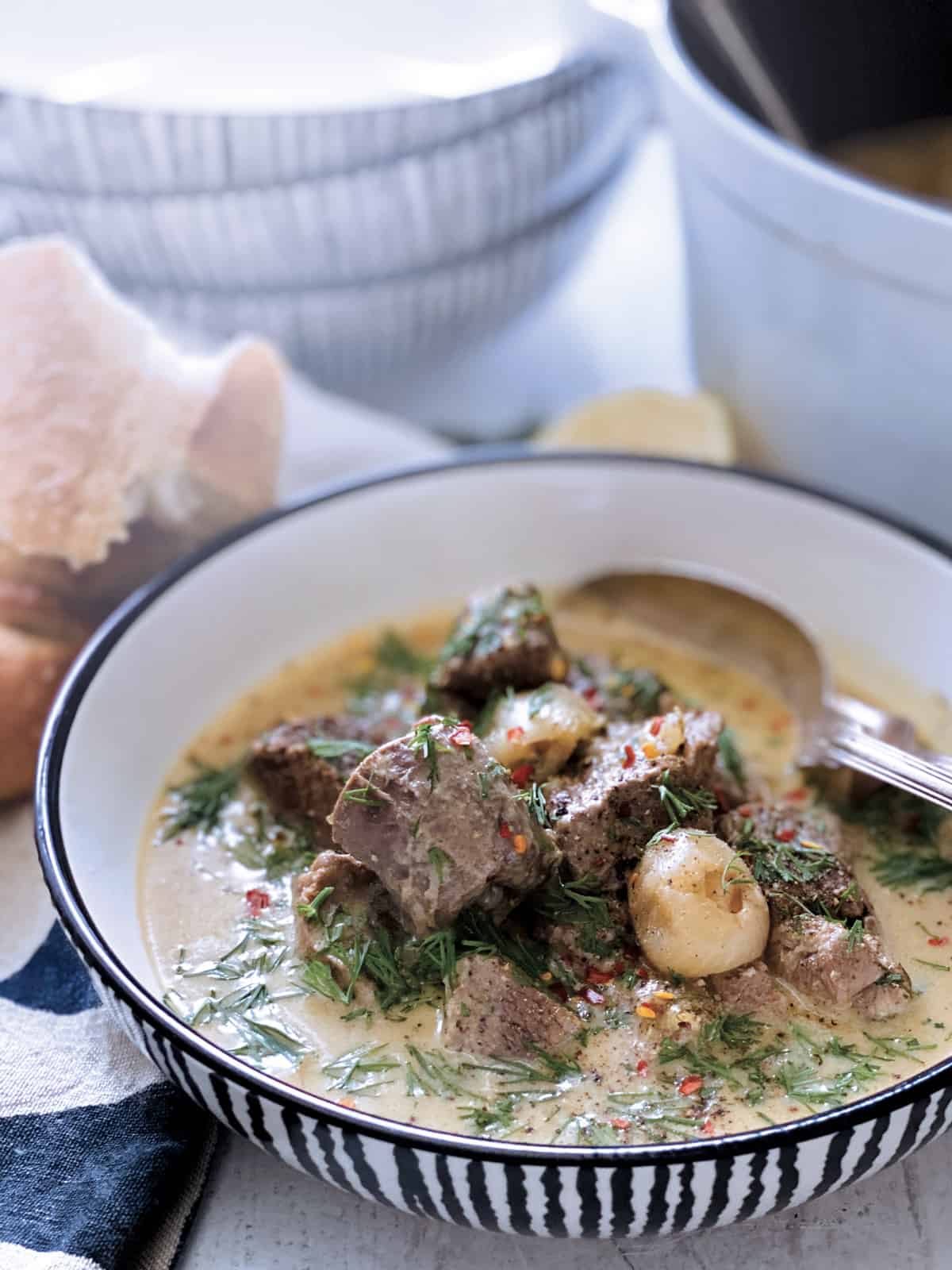 A plate with lamb stew avgolemono, a fork and spoon, half lemon at the back and a piece of bread on a white wooden table.