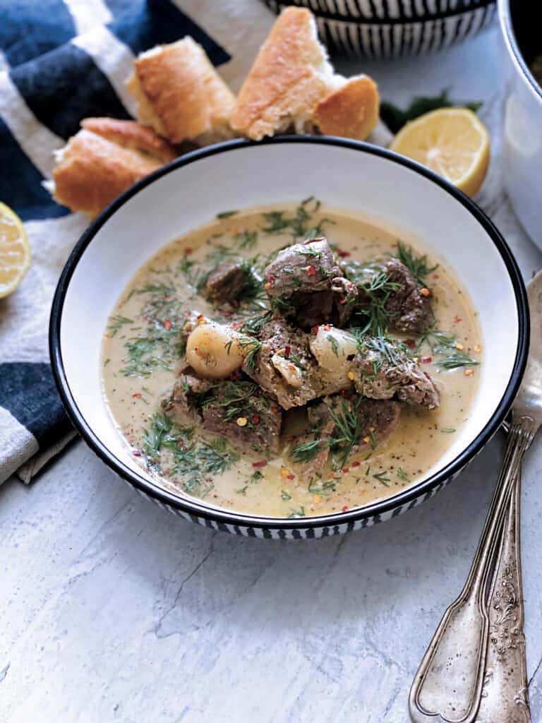 A plate with lamb stew avgolemono, a fork and spoon, half lemon at the back and a piece of bread on a white wooden table.