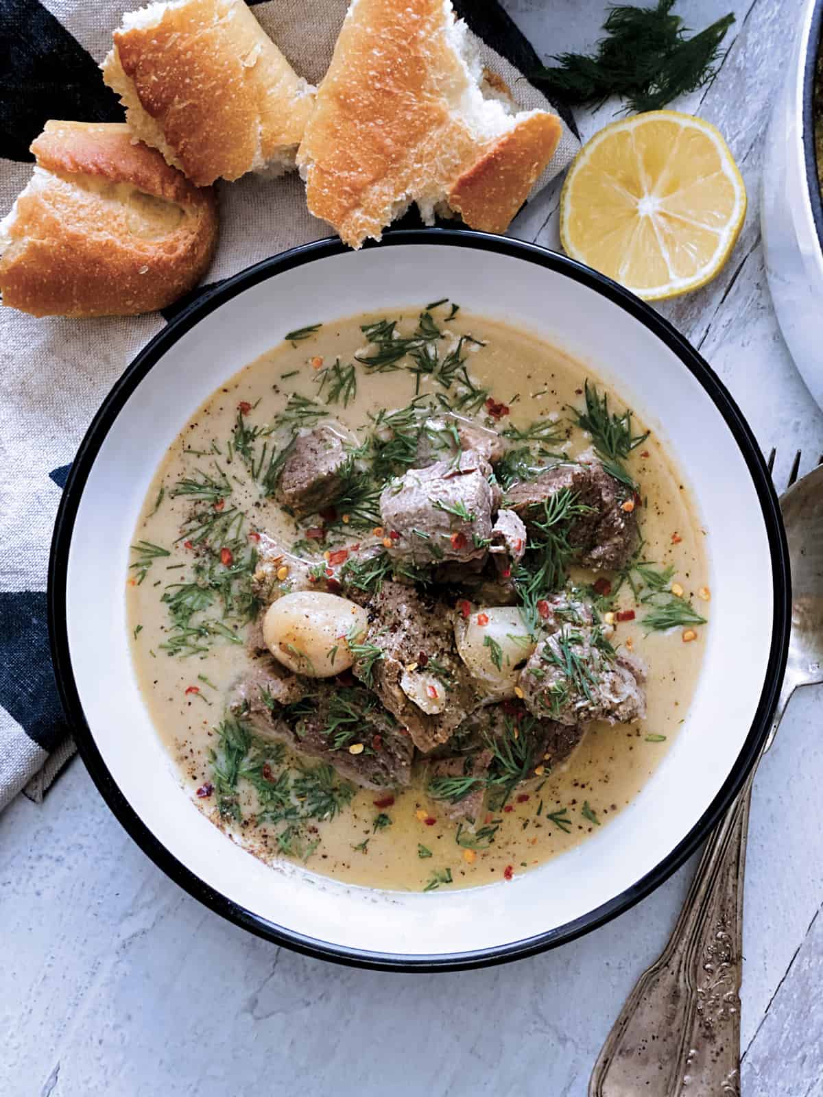 A plate with lamb stew avgolemono, a fork and spoon, half lemon at the back and a piece of bread on a white wooden table. 