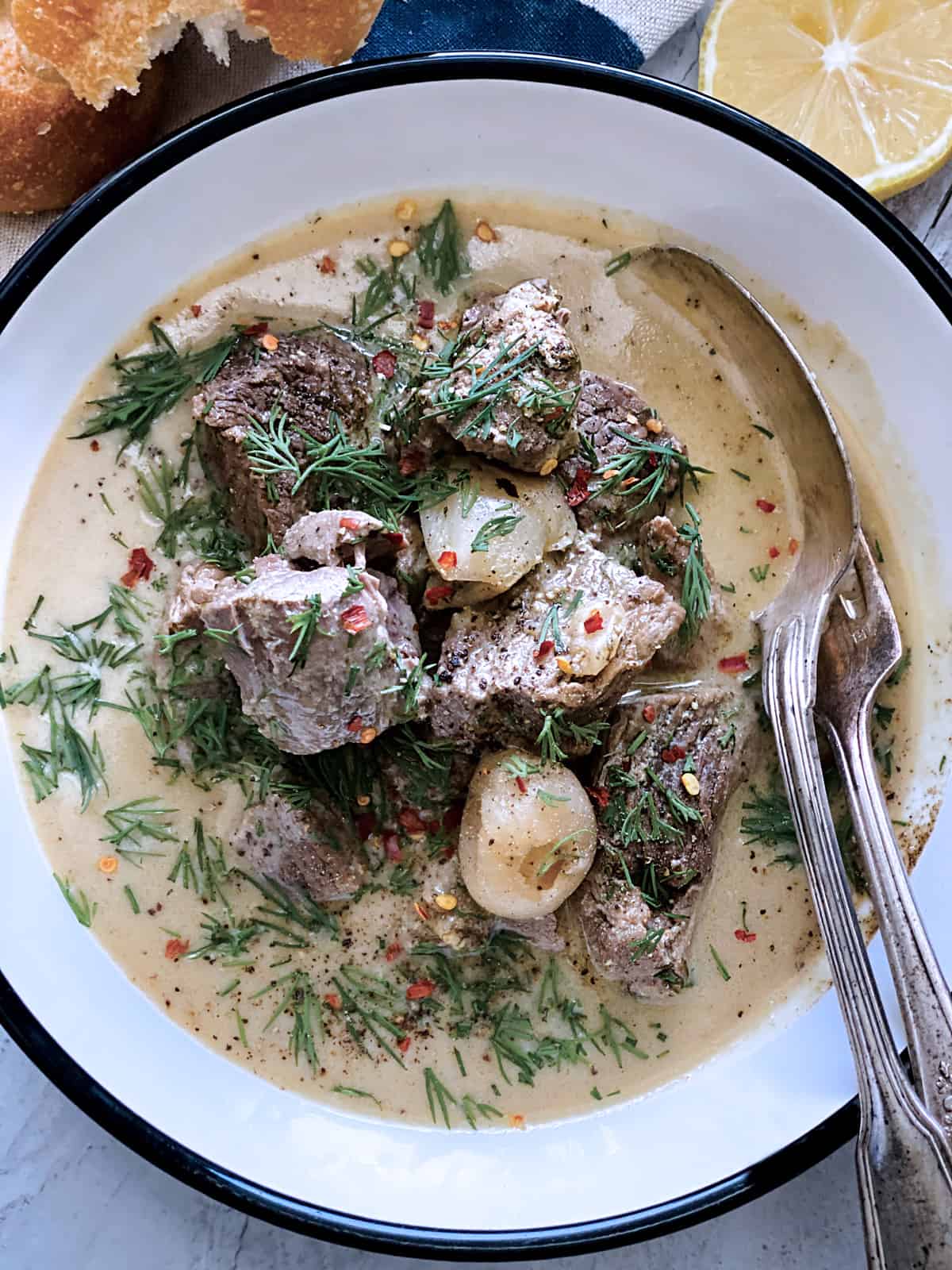 A plate with lamb stew avgolemono, a fork and spoon, half lemon at the back and a piece of bread on a white wooden table.