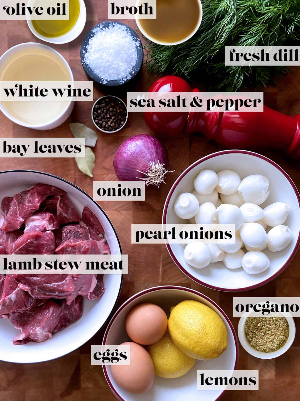 Ingredients for lamb stew. Small bowls with salt, olive oil, broth, pepper, white wine. A bunch of fresh dill, a pepper grinder, two bay leaves, an onion, a bowl with lamb stew cubes, a bowl with pears onions, a bowl with two eggs and two lemons and a small bowl with oregano, all on a butcher block. 