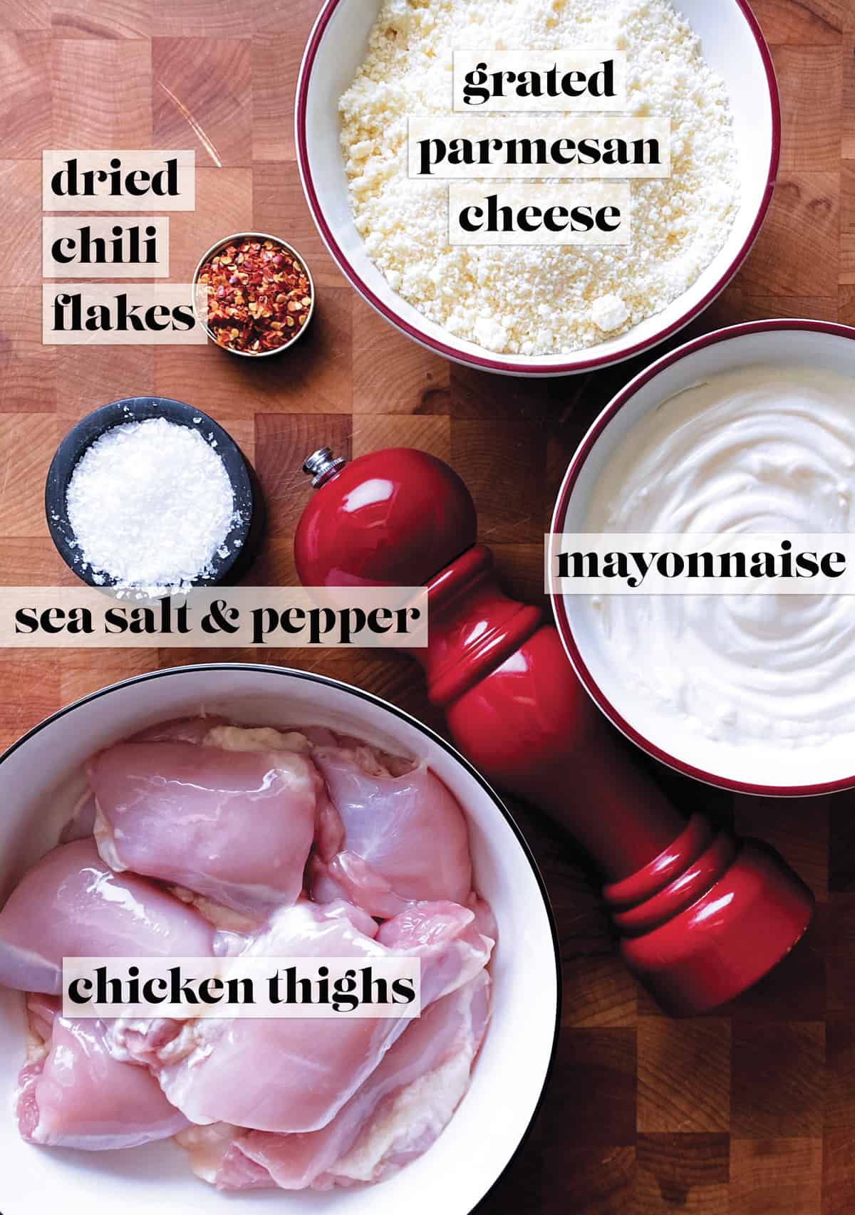 A bowl with grated parmesan cheese, a bowl with mayonnaise, a bowl with raw chicken thighs, a pepper mill, a small bowl with sea salt and a small bowl with chili flakes, all on a butcher block.