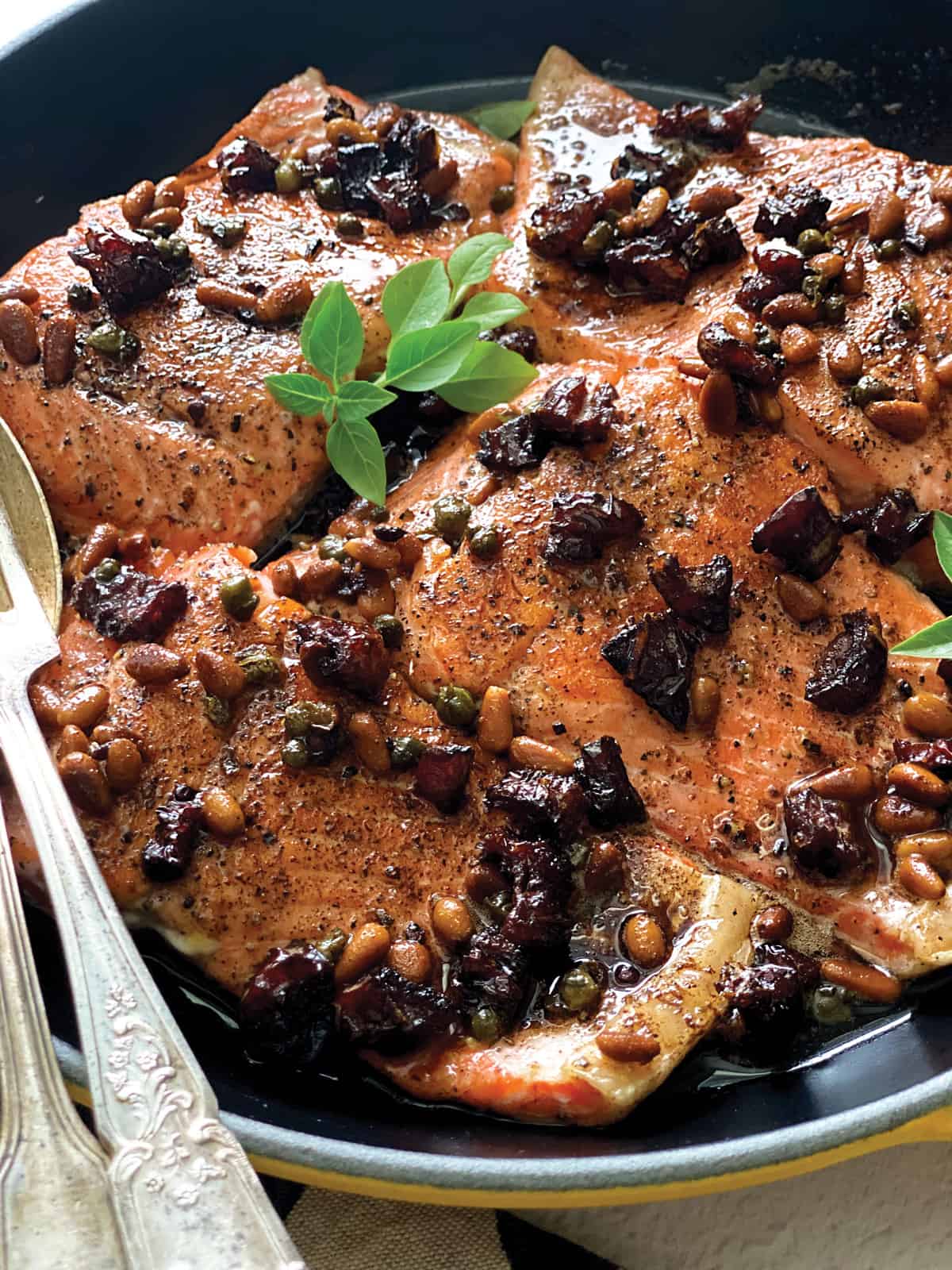 Brown butter salmon with capers and sundried tomatoes in a yellow pan with utencils.