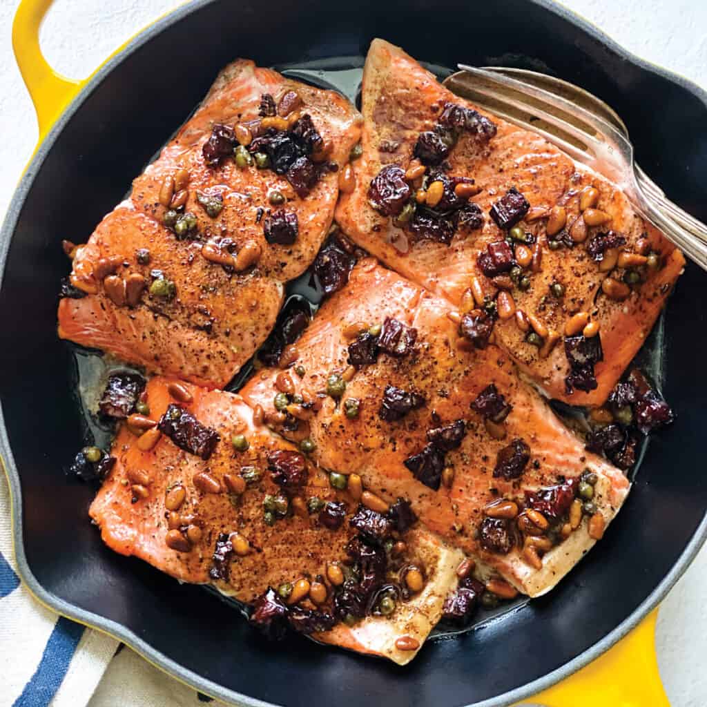 Brown butter salmon with capers and sundried tomatoes in a yellow pan with a cloth napkin on a white table.