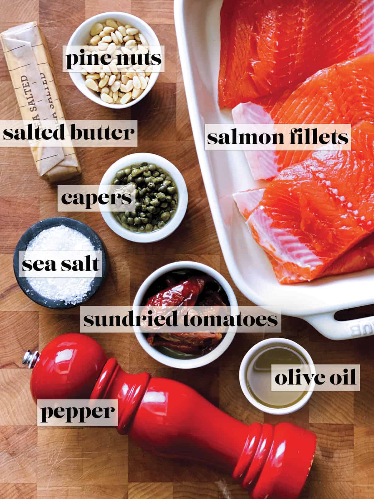 A baking pan with fresh salmon fillets, a pepper mill, a bowl with sundried tomatoes, a bowl with pine nuts, a bowl with capers a bowl with olive oil and a bowl with salt and a stick of butter.