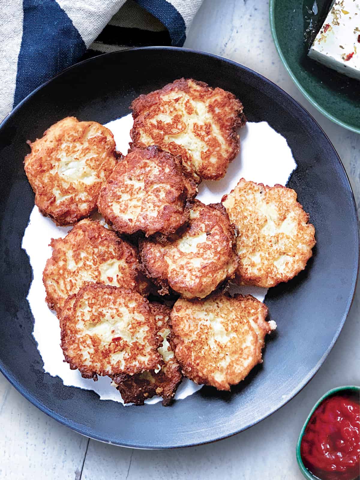 A plate with cauliflower fritters, a bowl with feta cheese and a small container with ketchup. A cloth napkin at the back.
