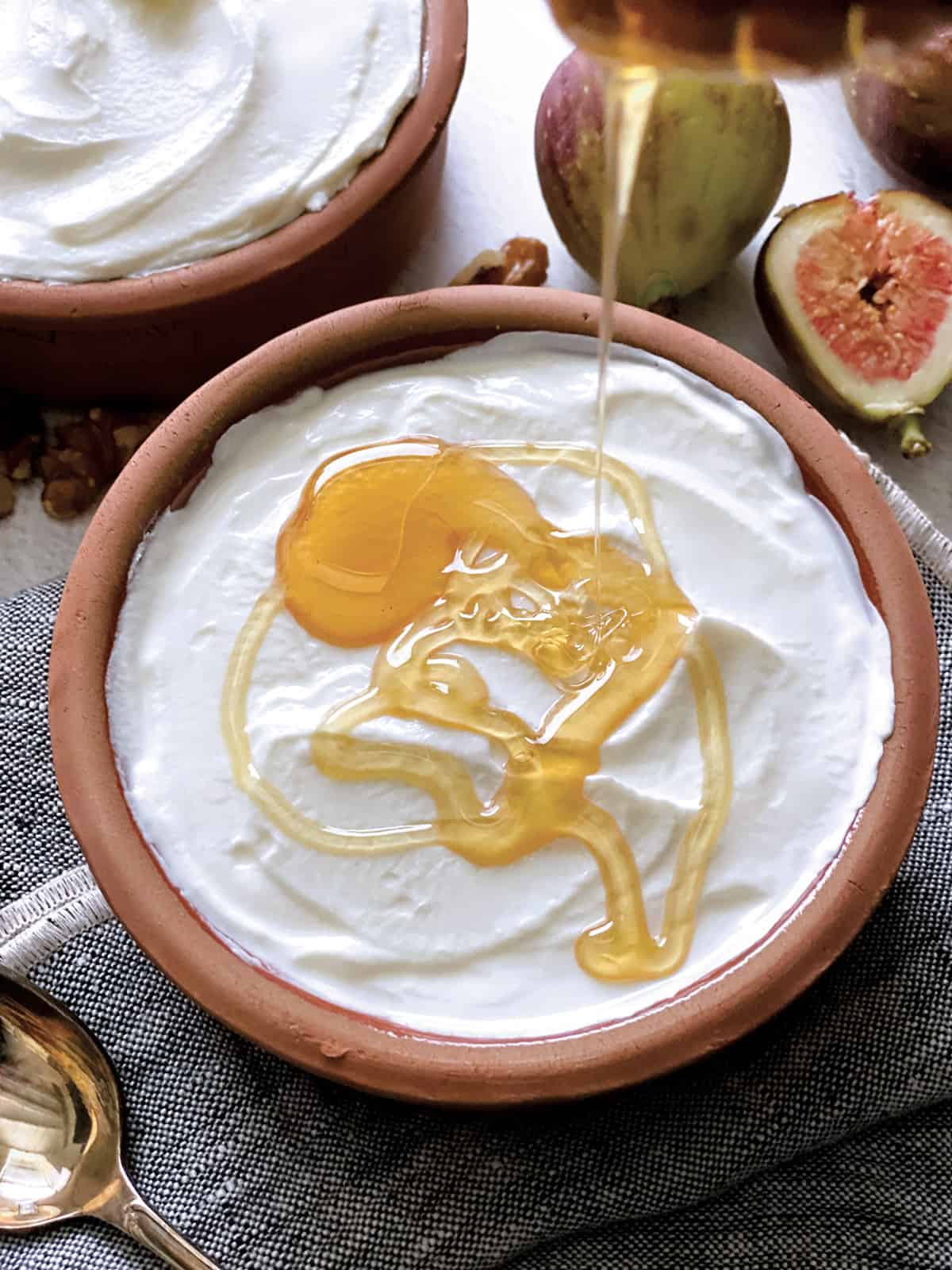 A bowl with yogurt, a honey dipper dripping honey, a spoon on a cloth napkin, fig halves, walnuts, and a detail of another bowl.