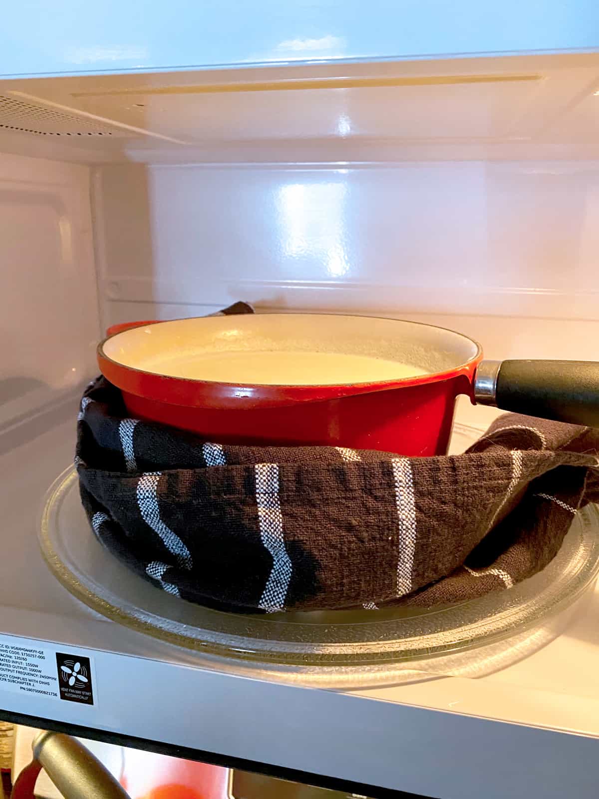 A red sauce pan with a towel wrapped around it in a microwave oven.