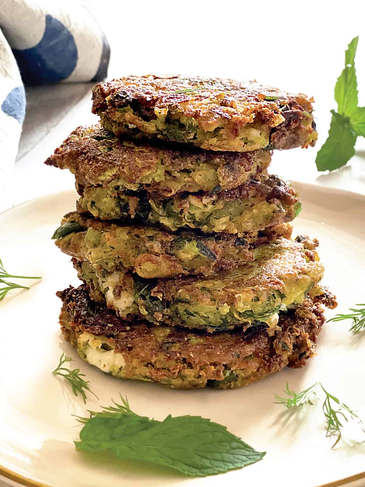 A plate with stacked fritters, at the back a napkin and some herbs.