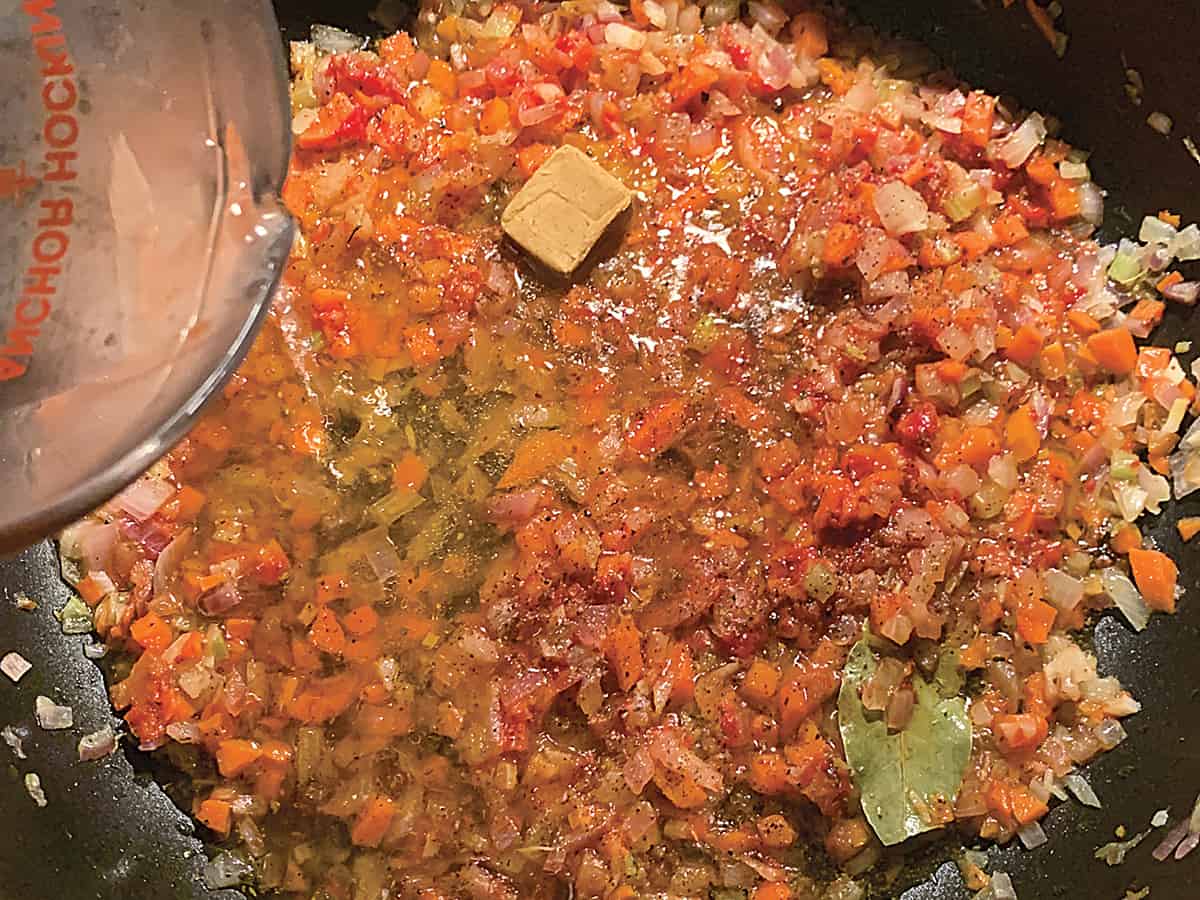 Diced onion and carrots with tomato paste, a bouillon cube and a bay leaf in olive oil and a jar pouring water.