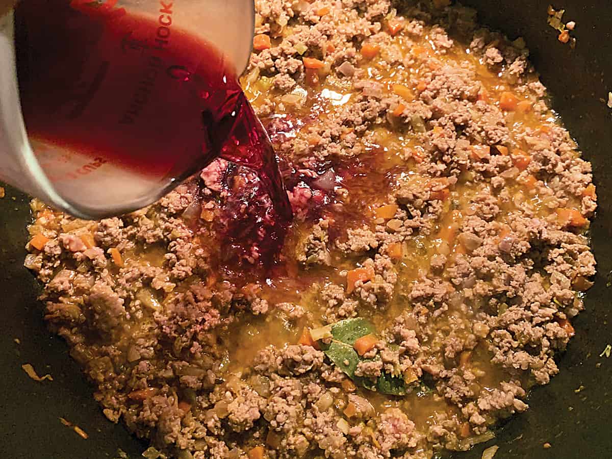 Diced vegetables and ground meat, a bouillon cube and a bay leaf in olive oil and a jar pouring wine.