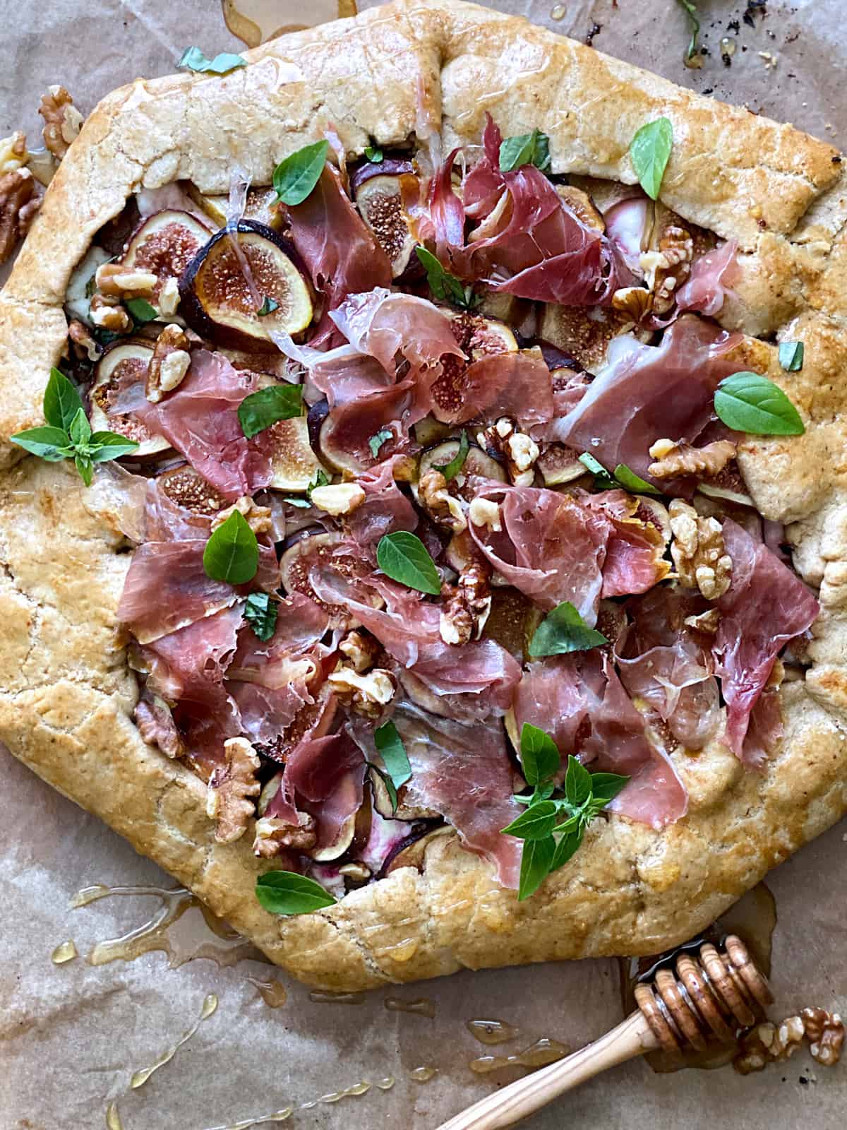 A savory galette with figs and prosciutto and a honey dipper on parchment paper.