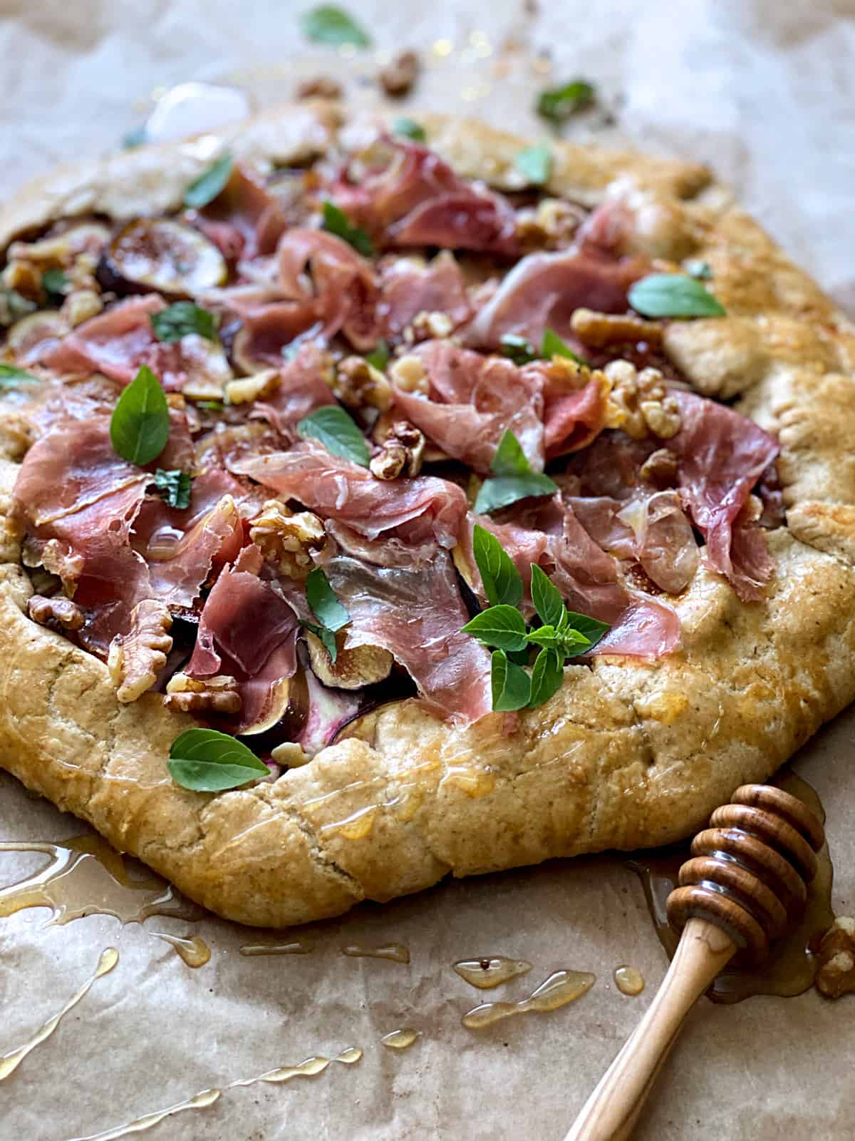 A savory galette with figs and prosciutto and a honey dipper on parchment paper.