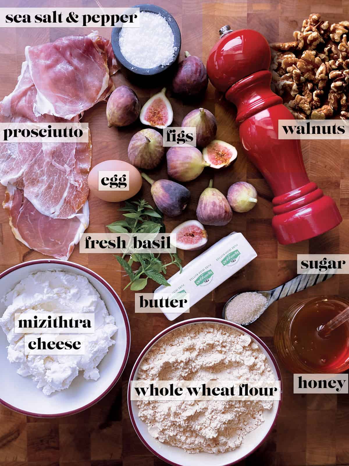 Slices of prosciutto, an egg, figs, a container with salt, a pepper mill, some walnuts, fresh basil, a spoon with sugar, a stick of butter, a bowl with cheese, a bowl with flour and a pot of honey.