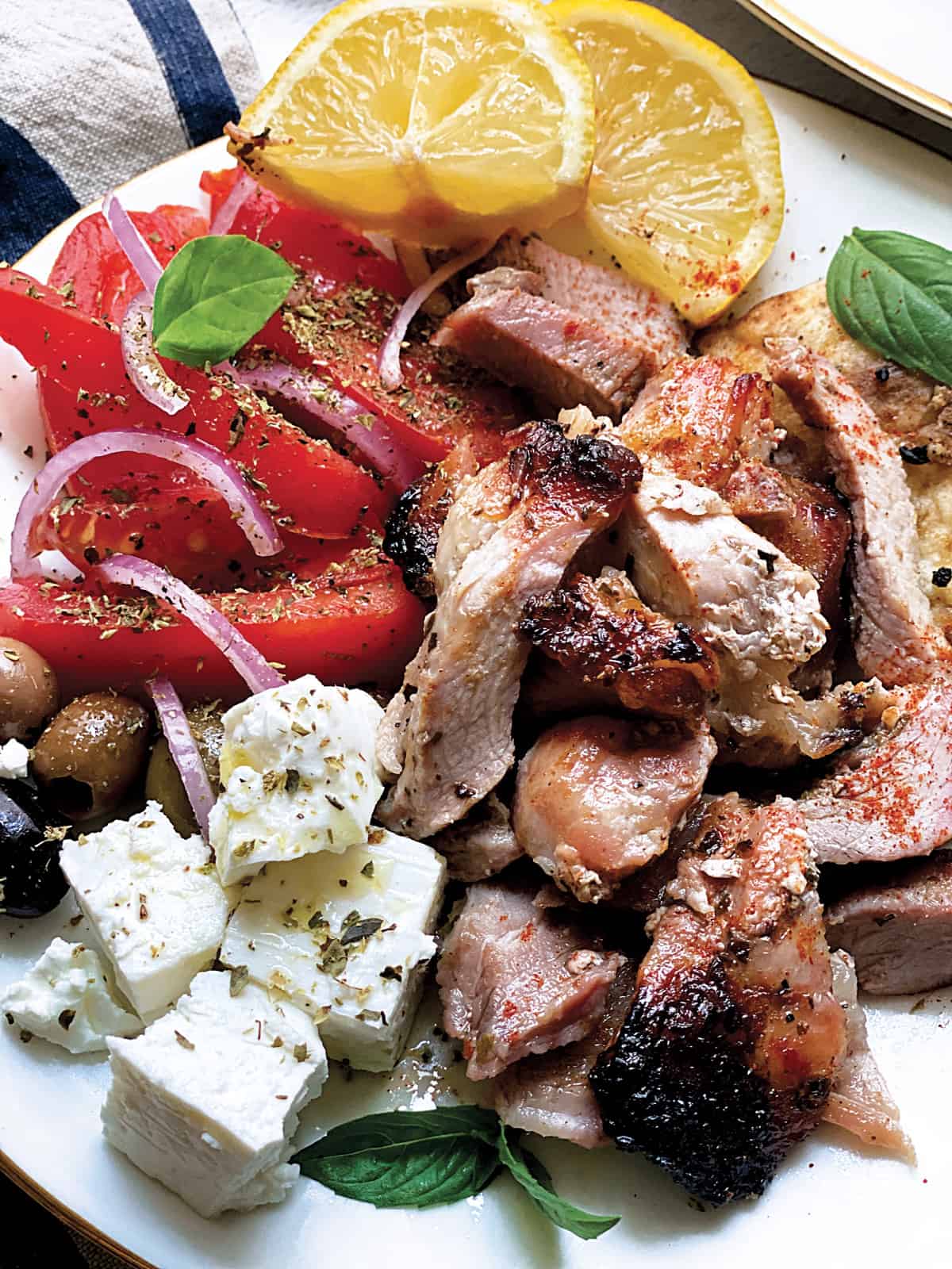 A plate with sliced tomatoes and onions, olives, feta cheese cubes, pieces of gyro, lemon wedges and pitas.