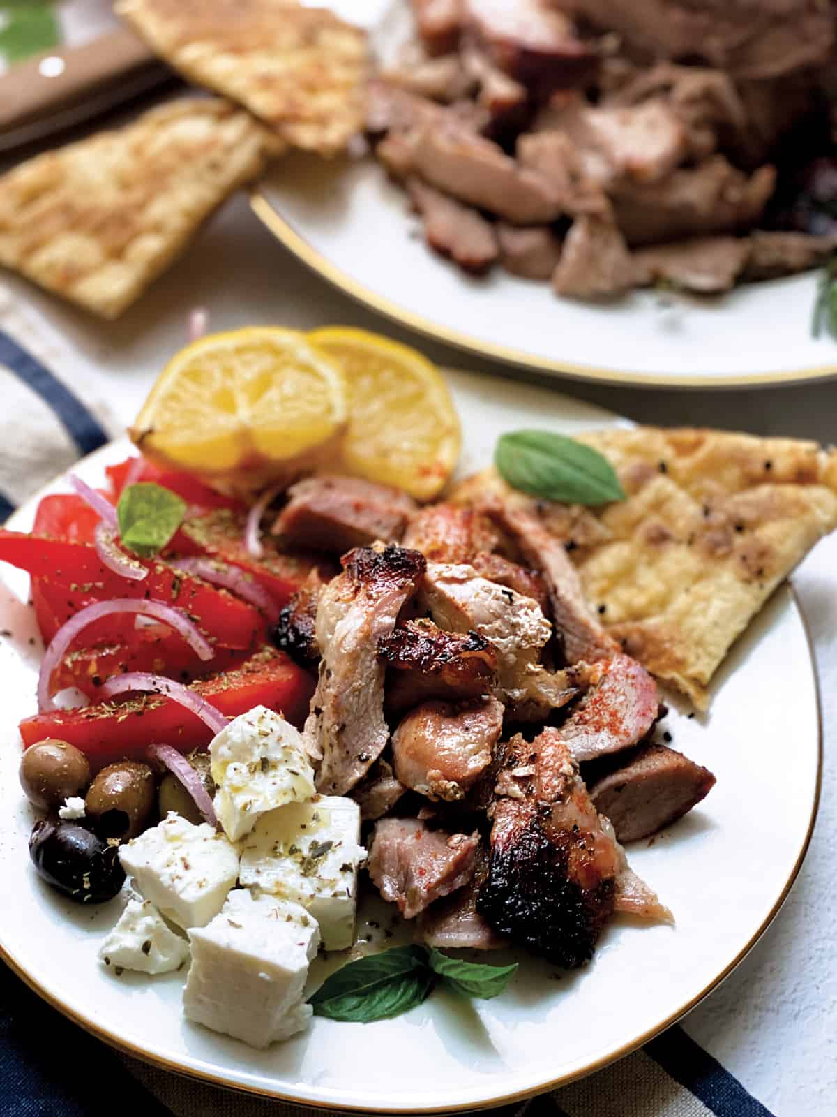 A plate with sliced tomatoes and onions, olives, feta cheese cubes, pieces of greek gyro lemon wedges and pitas.