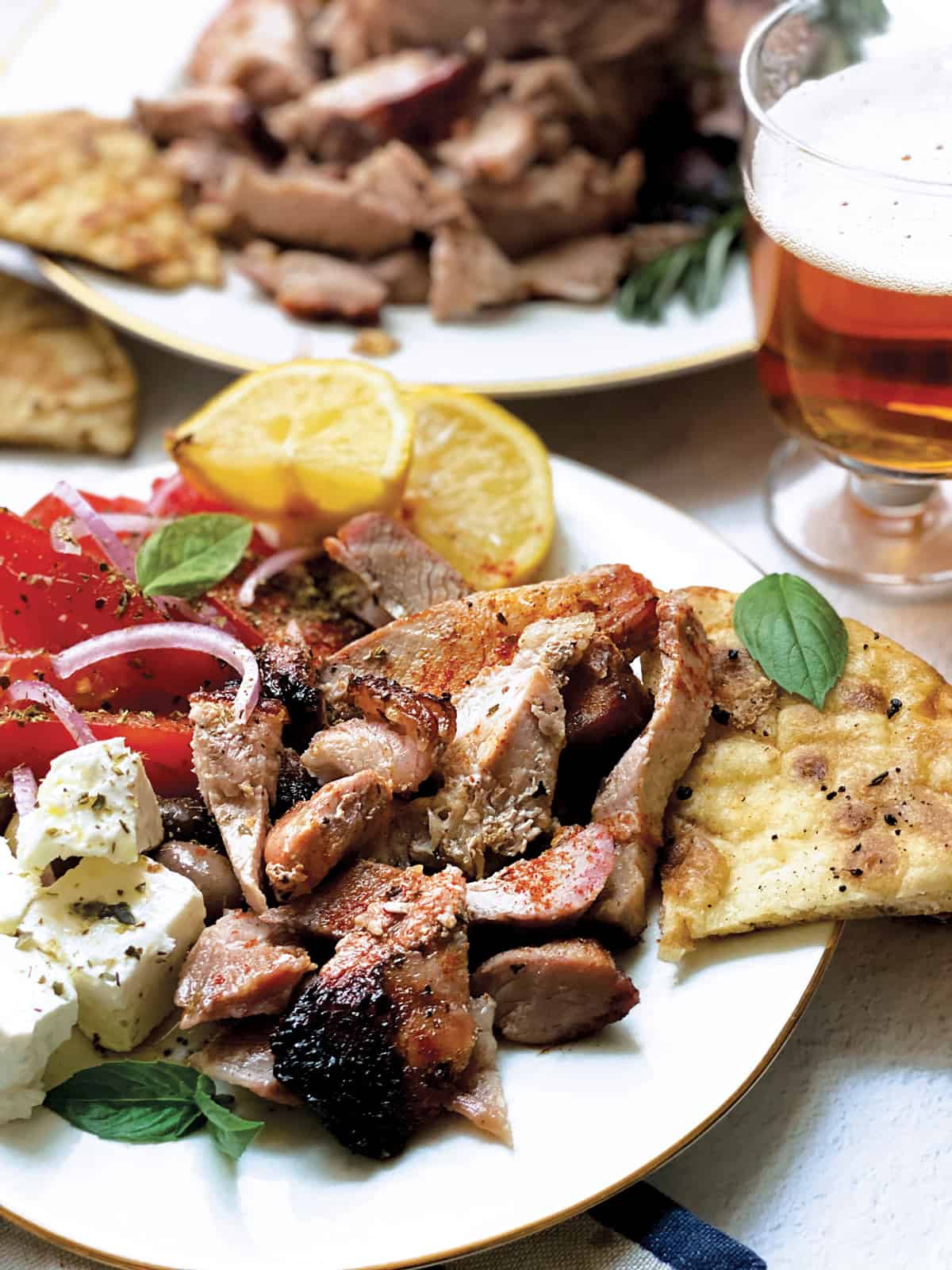 A plate with sliced tomatoes and onions, olives, feta cheese cubes, pieces of greek gyro lemon wedges and pitas.