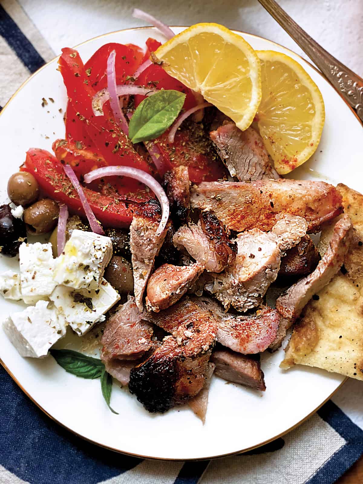 A plate with sliced tomatoes and onions, olives, feta cheese cubes, pieces of greek gyro lemon wedges and pitas.