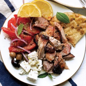 A plate with sliced tomatoes and onions, olives, feta cheese cubes, pieces of greek gyro lemon wedges and pitas.