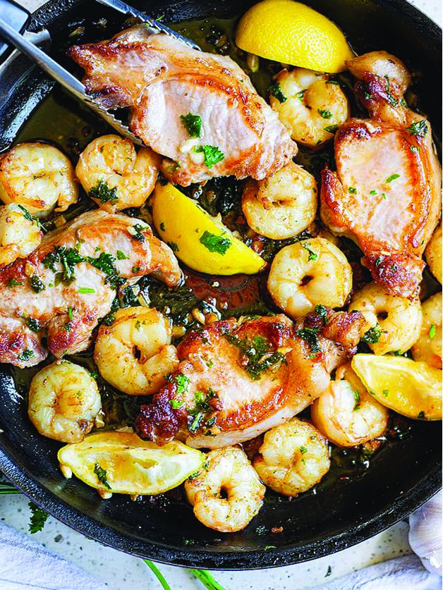 A skillet with pork chops , lemon wedges and shrimp.