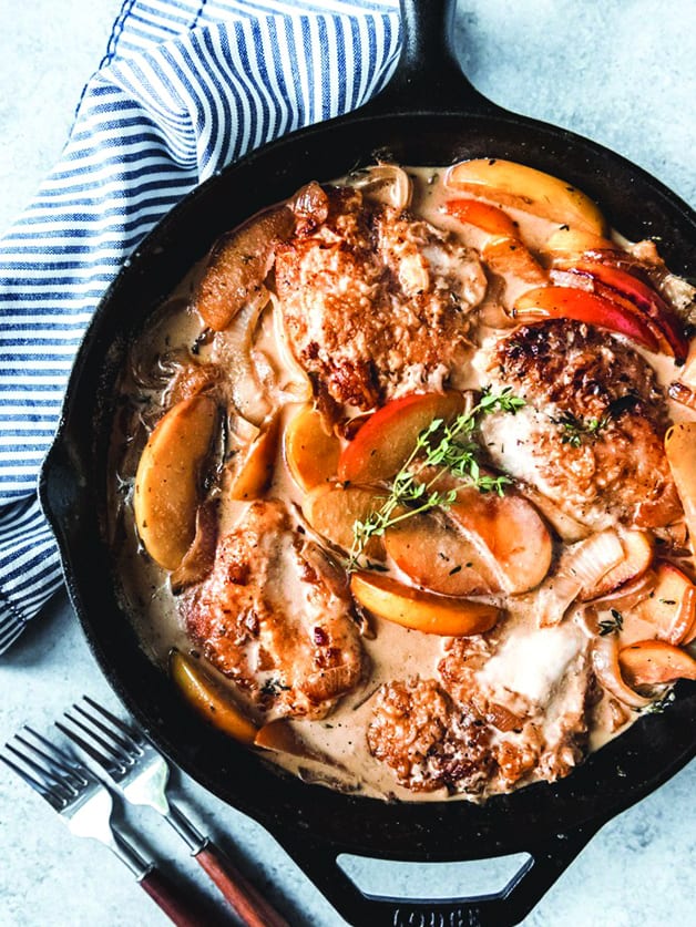 A skillet with chicken, apple slices, creamy sauce and herbs on a cloth napkin.