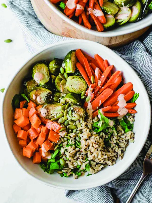 A bowl with roasted brussels sprouts, carrots, sweet potatoes, fresh greens, brown rice, quinoa, and pepitas.