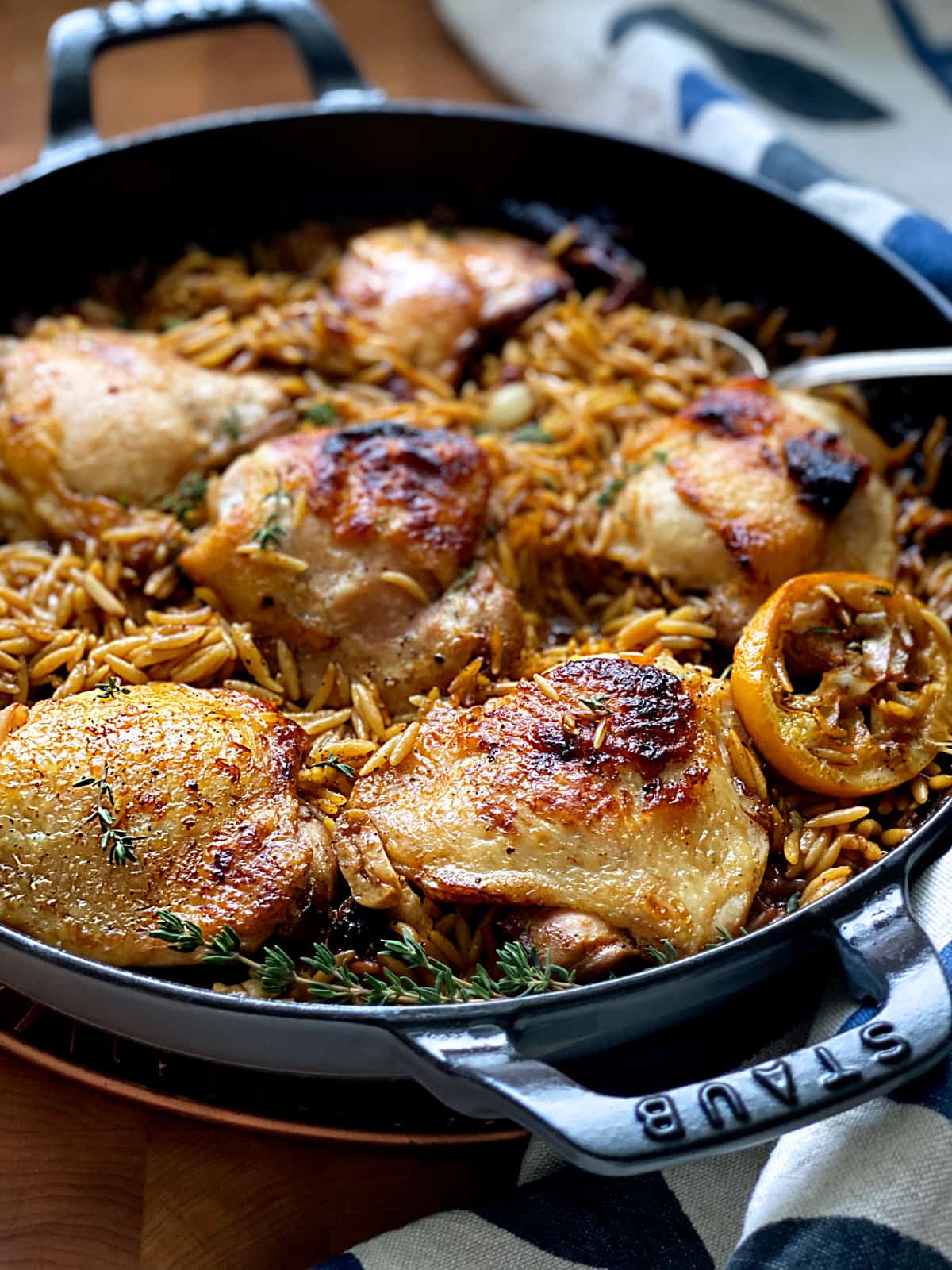 Butter milk chicken thighs with orzo in pan with a spoon, and a kitchen towel on a wooden table.