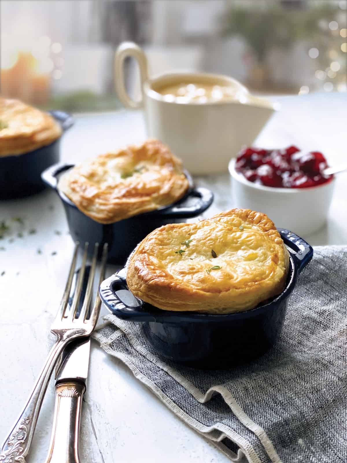 Two mini pot pies a fork and knife and a bowl with cranberry sauce and a gravy boat at the back in a holiday environment.