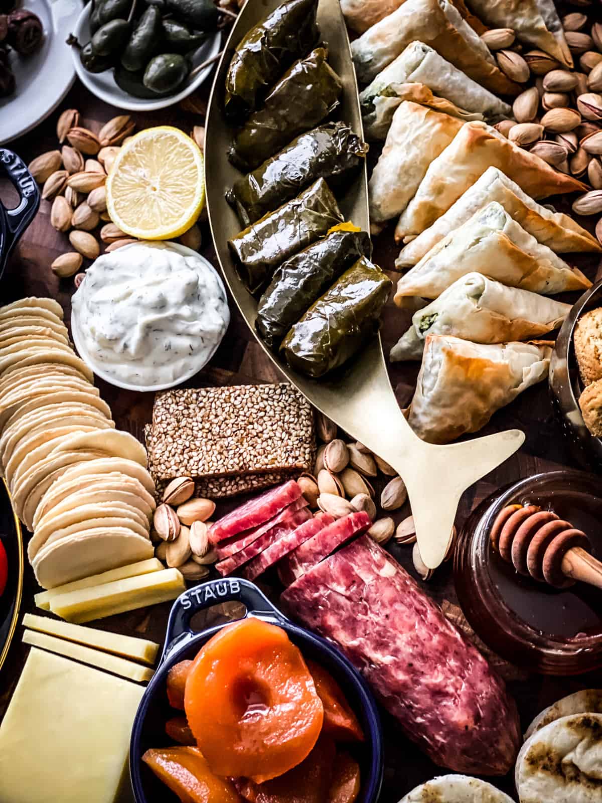 Anchovies on a plate, large capers, , some pistachios, tomato and cucumbers and feta in a bowl, cheese and spinach triangles, a small pot with gigantes beans, a bowl with olives.