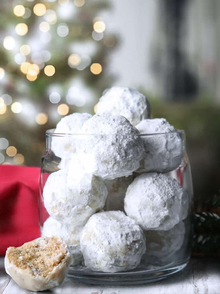 A glass vase with greek Christmas cookies, kourabiedes, a red napkin and a tree with lights at the back.