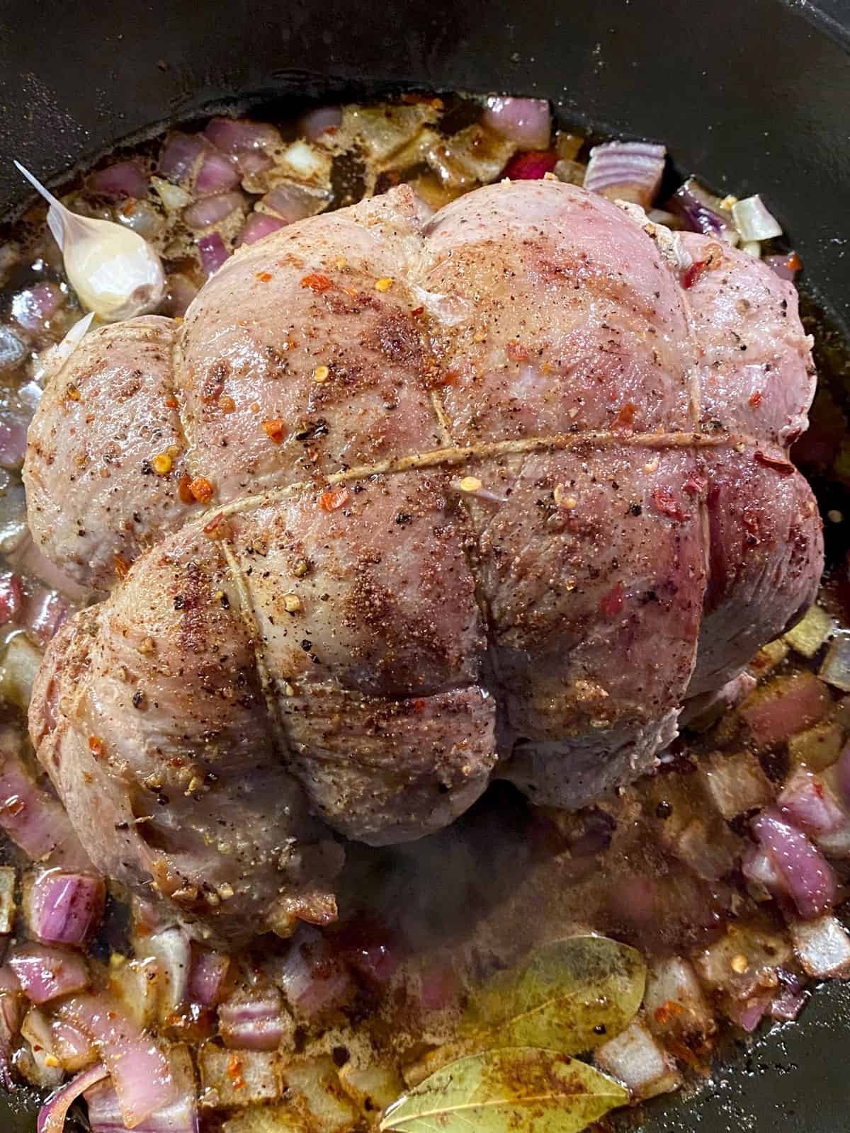A pork shoulder roast tied with twine  in a skillet with diced onions and bay leaves.