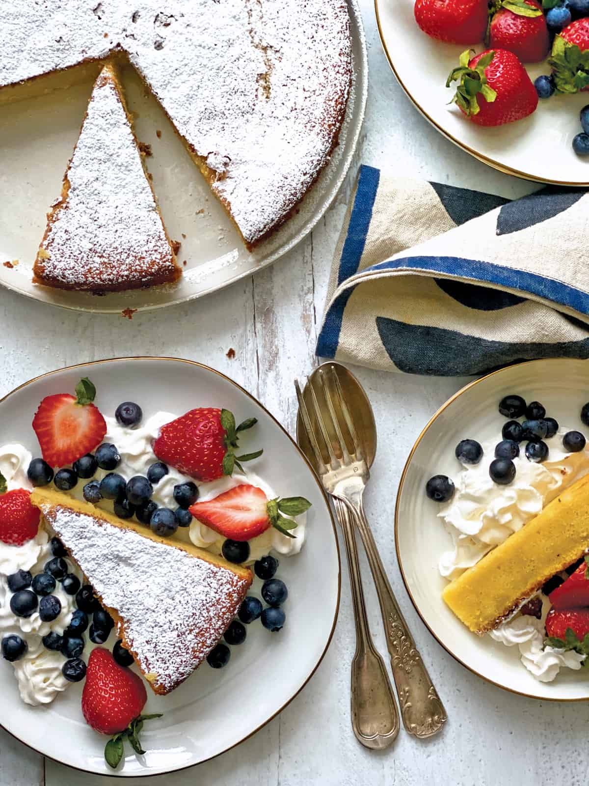 A piece of Meyer lemon olive oil cake with whipped cream and berries on two plates, at the back a cake on a platter.