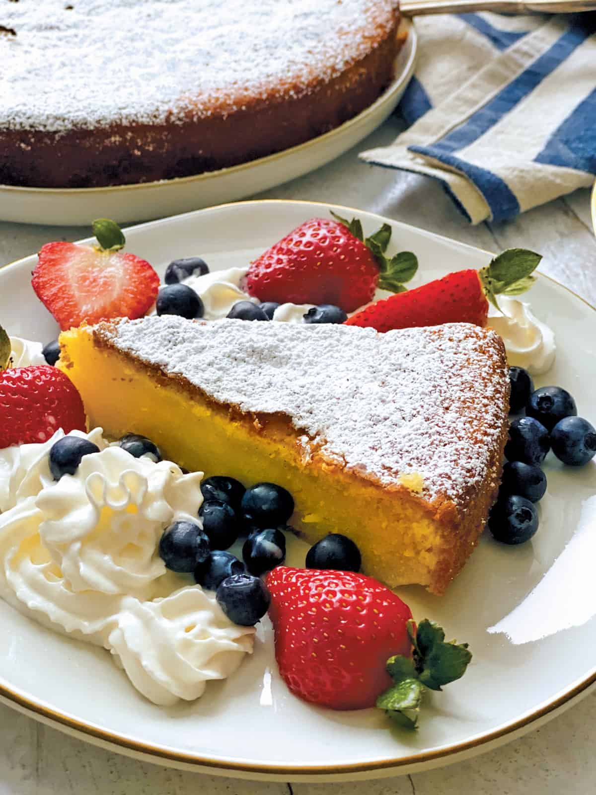 A piece of Meyer lemon olive oil cake with whipped cream and berries on a plate, at the back a cake on a plater.