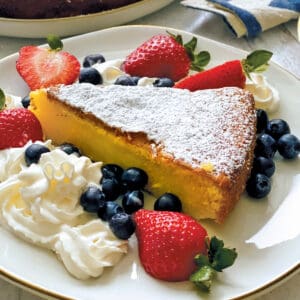 A piece of Meyer lemon olive oil cake with whipped cream and berries on a plate, at the back a cake on a platter.