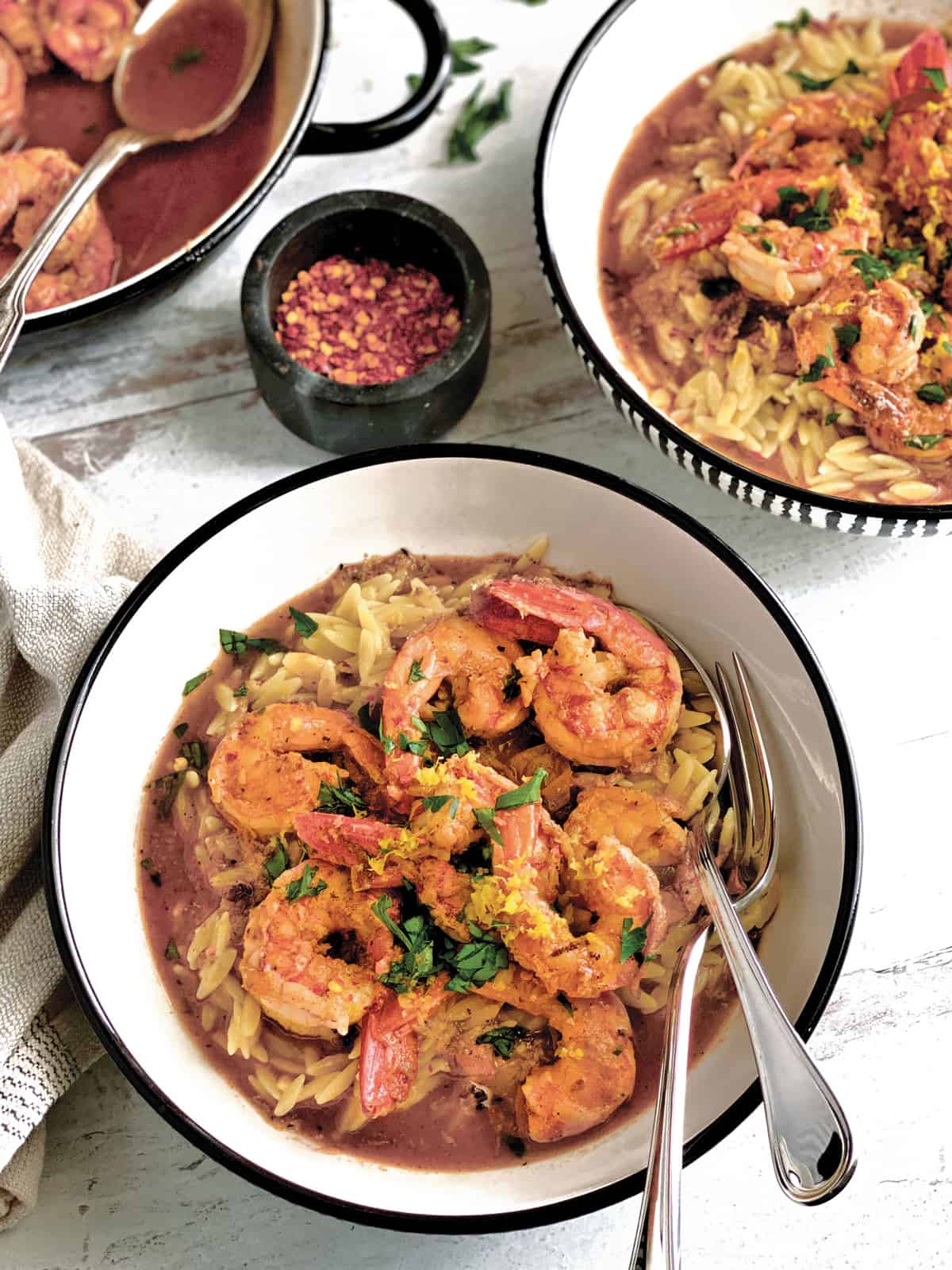 Two bowls with shrimp orzo in lemon butter sauce and utensils. Side view of a pan with shrimp and sauce and a spoon.