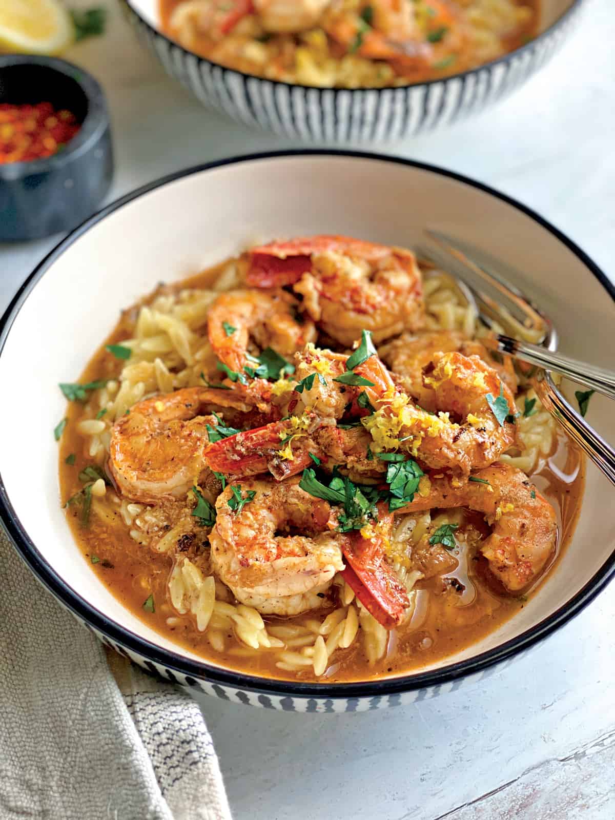 A bowl with shrimp orzo in lemon butter sauce and utensils, a cloth napkin under the bowl.