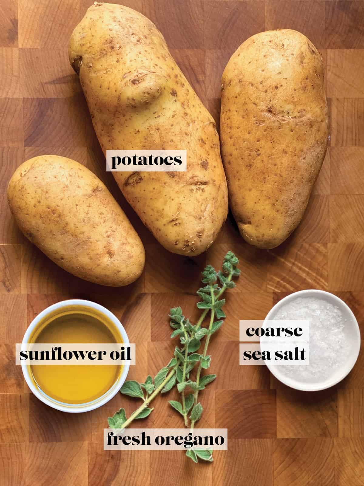 Three potatoes, small containers with sunflower oil, and sea salt, and some fresh oregano on a butcher block.